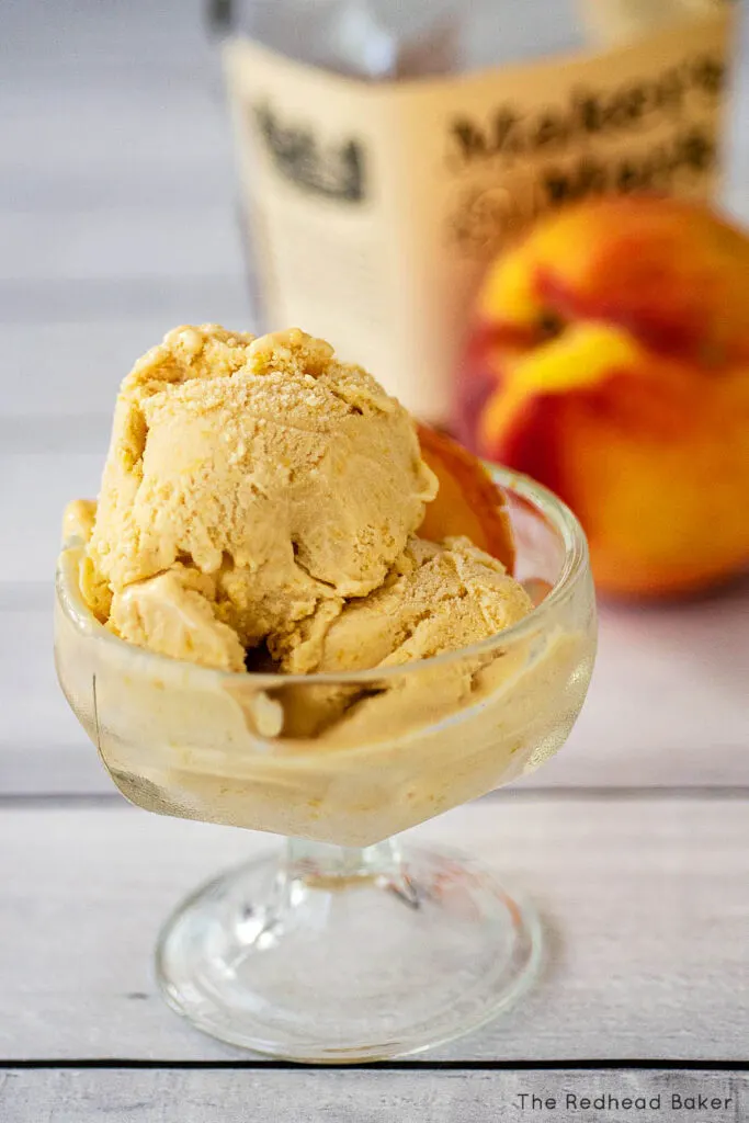 Two scoops of ice cream with a peach slice in front of a bottle of bourbon and a fresh peach