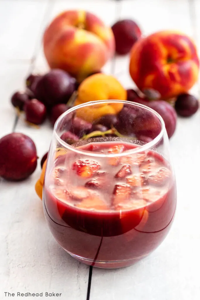 A close-up of a glass of grilled stone-fruit sangria with various stone fruits in the background