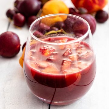 A close-up of a glass of grilled stone-fruit sangria with various stone fruits in the background