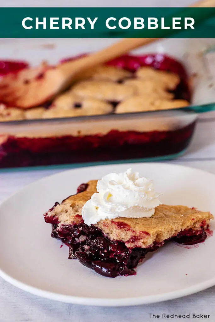 A portion of cherry cobbler on a white plate topped with whipped cream, in front of the remaining cobbler in a glass baking dish