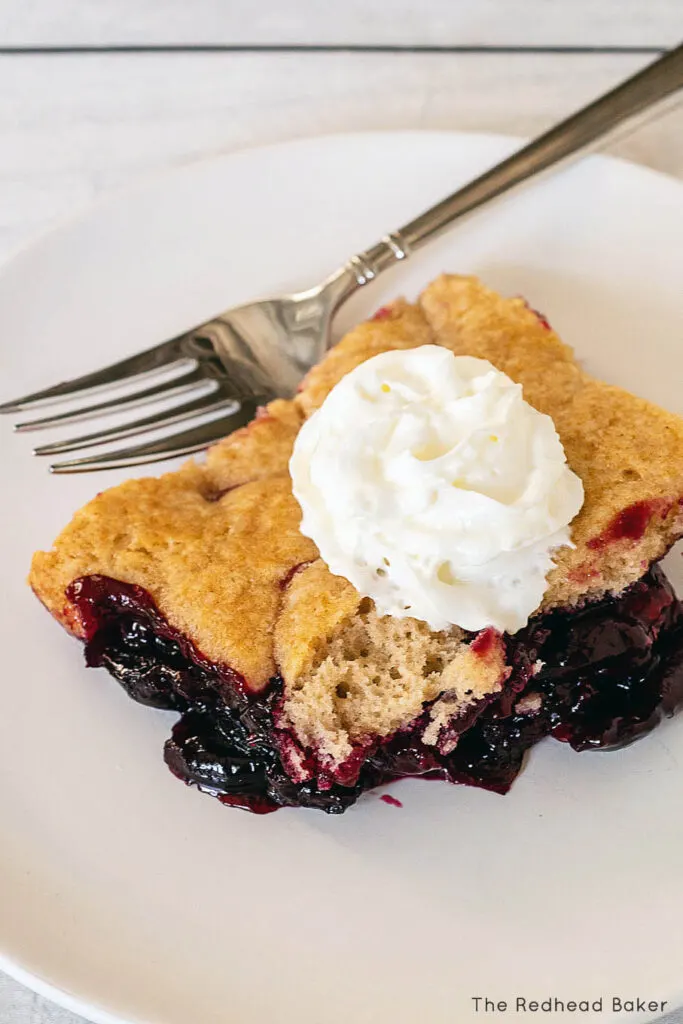 An overhead view of a fork and a portion of cherry cobbler on a white plate, topped with whipped cream