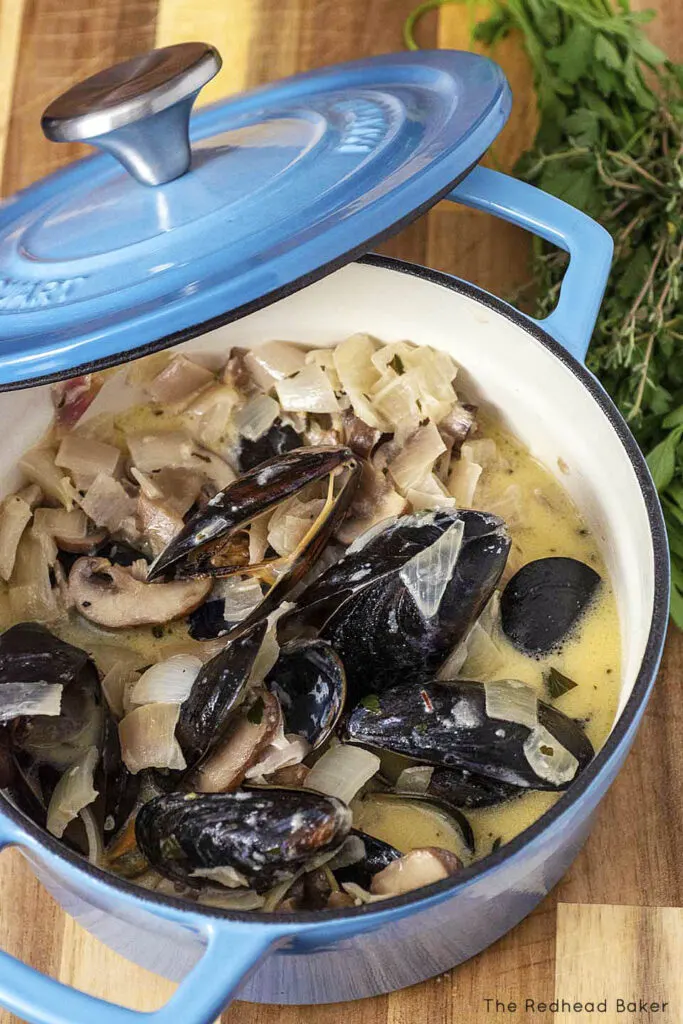 Looking down into a mini Dutch oven of Normandy-style mussels in cider sauce, with a handful of parsley and thyme behind the pot