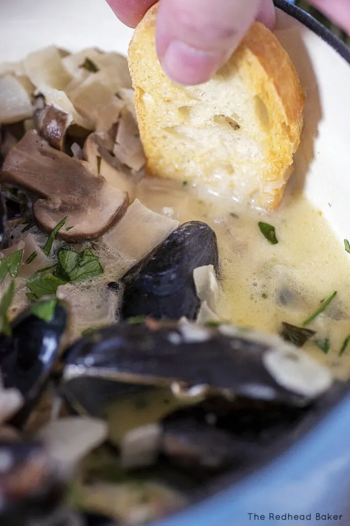 A slice of toasted baguette being dipped into the cider sauce of the mussels