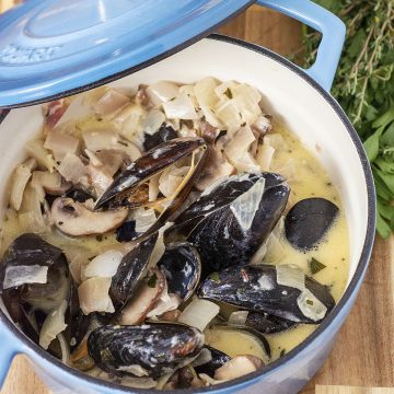 Looking down into a mini Dutch oven of Normandy-style mussels in cider sauce, with a handful of parsley and thyme behind the pot