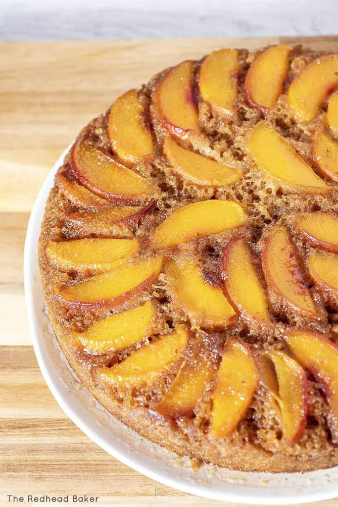 A baked peach upside-down cake on a white serving plate. The plate is sitting on a wooden cutting board.