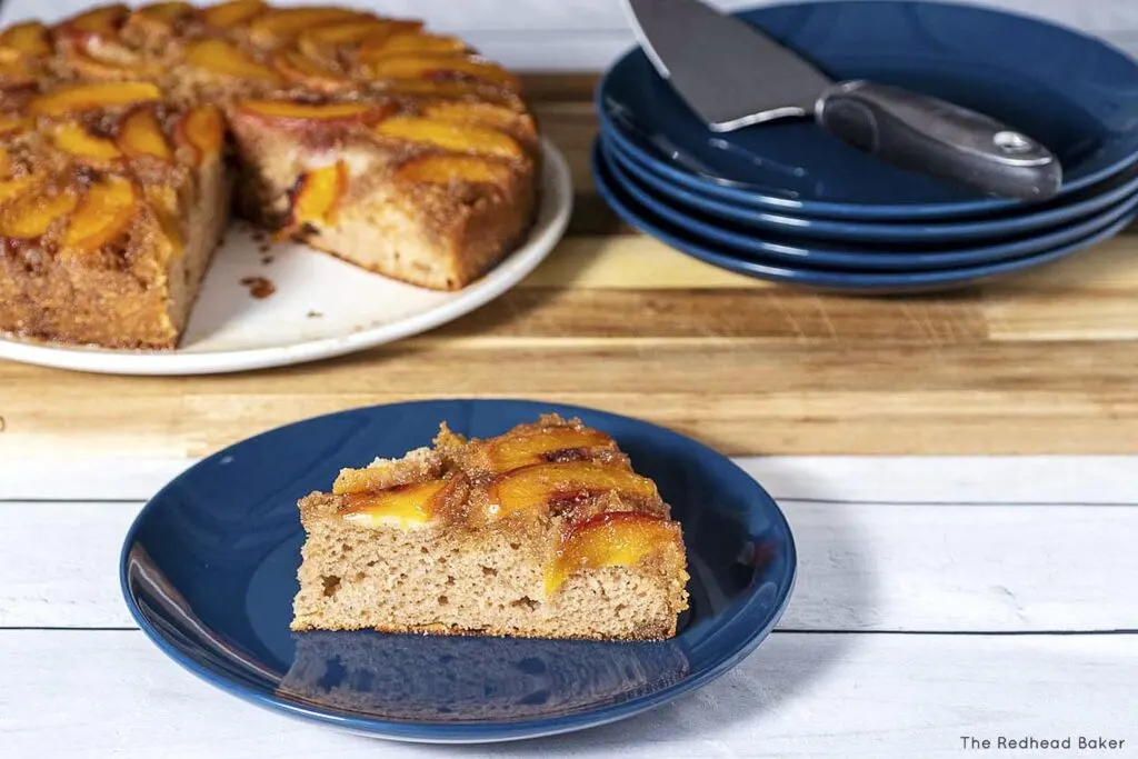 A slice of peach upside-down cake on a blue plate in front of the rest of the cake