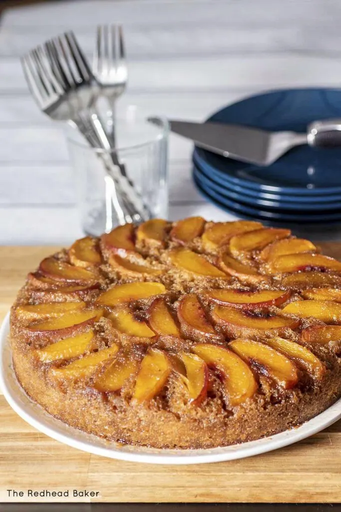 An upside-down cake on a white serving plate, in front of a stack of dessert plates and several forks standing upright in a glass