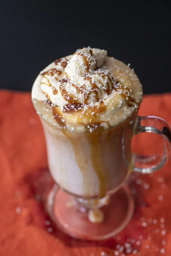 An overflowing mug of pumpkin white hot chocolate, topped with caramel whipped cream, caramel sauce drizzle and white chocolate shavings