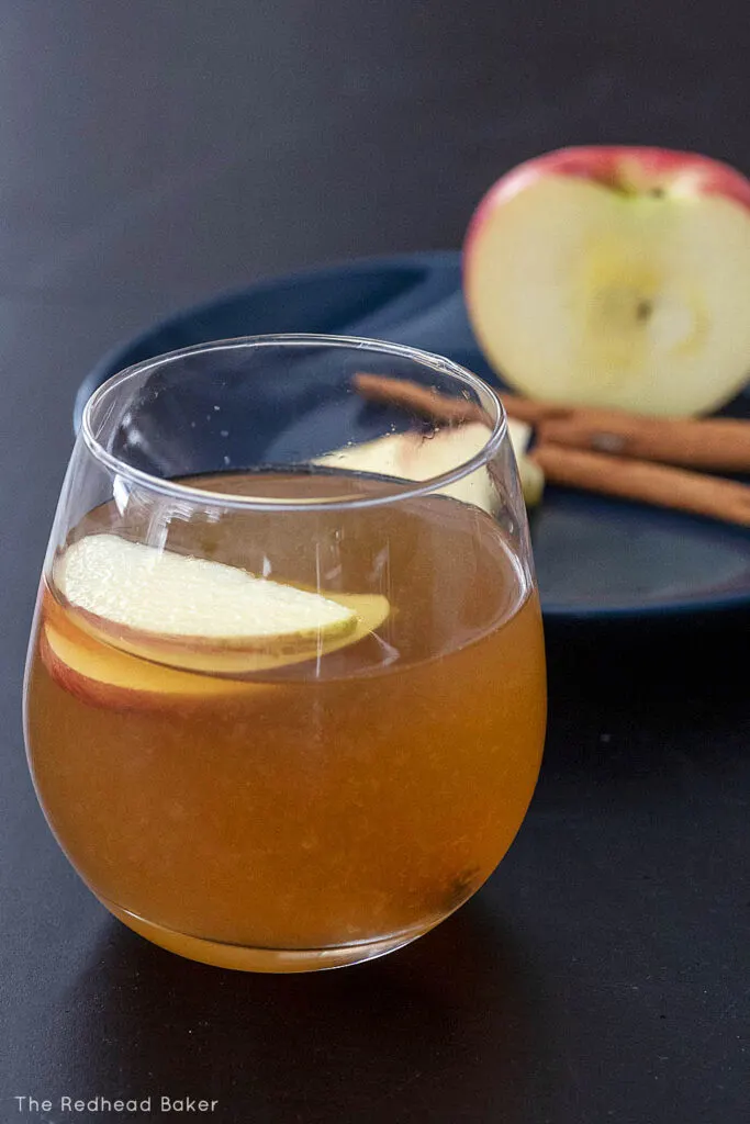 A glass of whiskey sour in front of a blue plate with apple slices, half an apple and cinnamon sticks