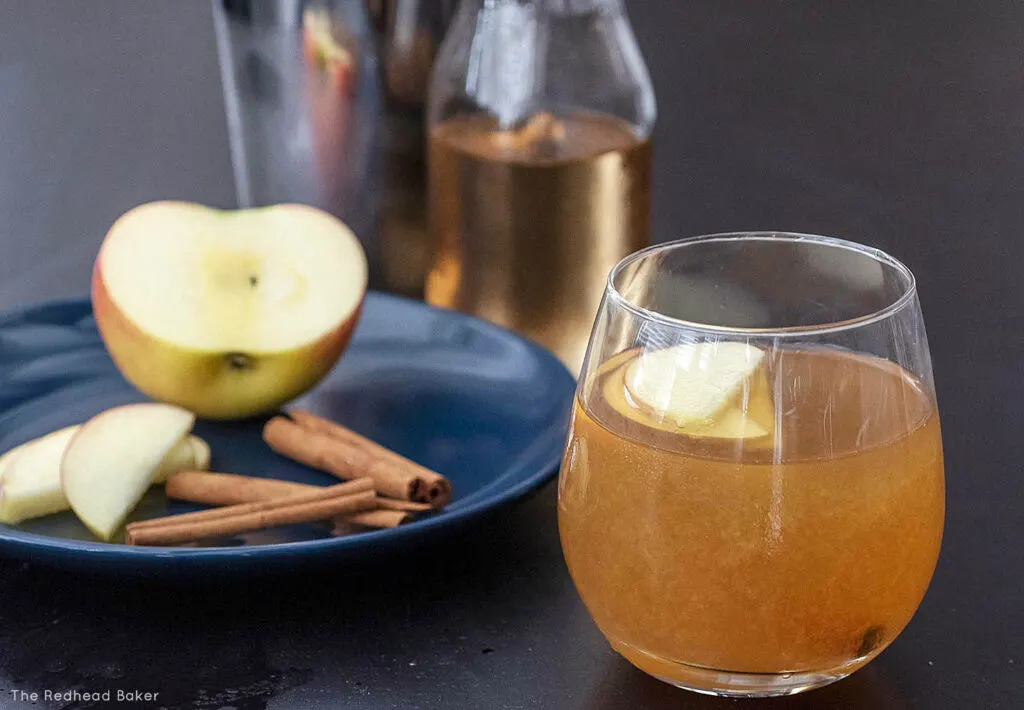 A cinnamon cider whiskey sour cocktail in front of a plate of sliced apple and cinnamon sticks, a cocktail shaker and a bottle of cinnamon syrup