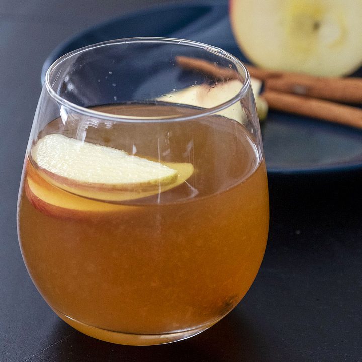A glass of whiskey sour in front of a blue plate with apple slices, half an apple and cinnamon sticks