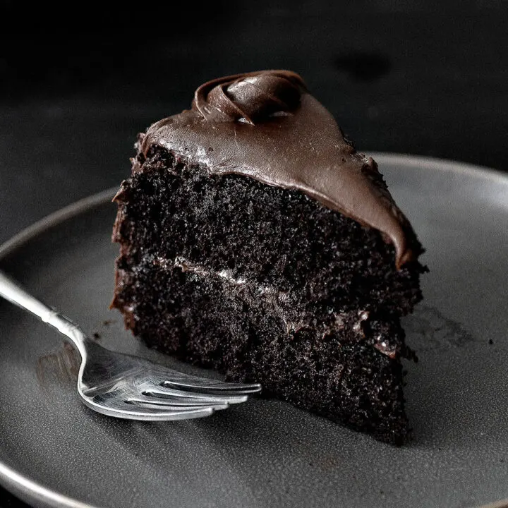 A slice of chocolate layer cake on a dark gray plate with a fork