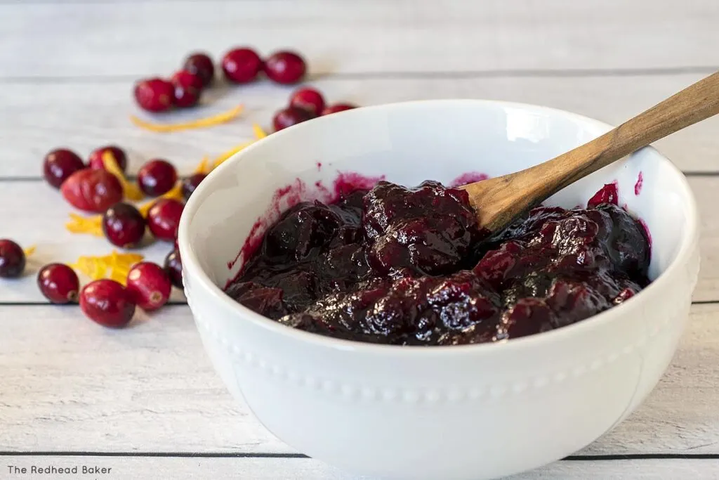 A bowl of cherry cranberry sauce in front of fresh cranberries and orange zest