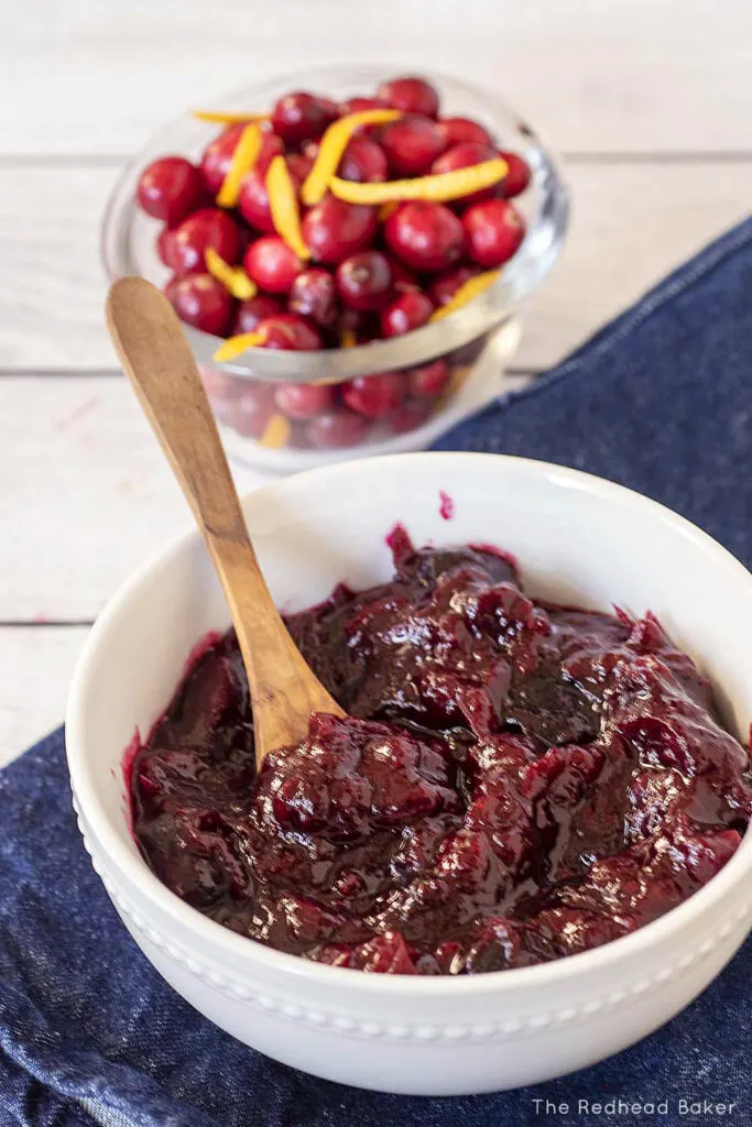 A white bowl filled with cherry cranberry sauce in front of a clear bowl filled with fresh cranberries and orange zest