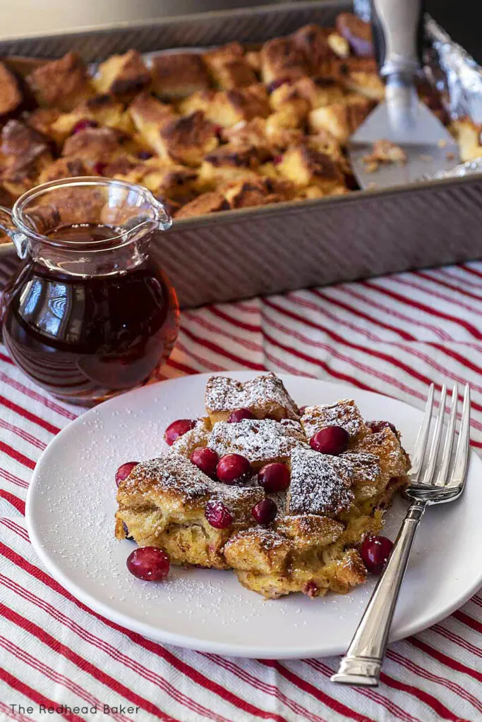 A plate of cranberry eggnog French toast bake in front of a baking dish and a container of maple syrup