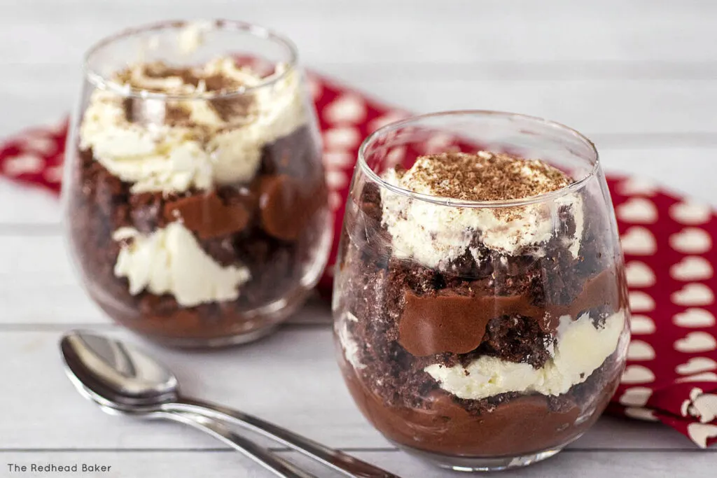 Two glasses of trifle in front of a red tea towel with hearts and two spoons