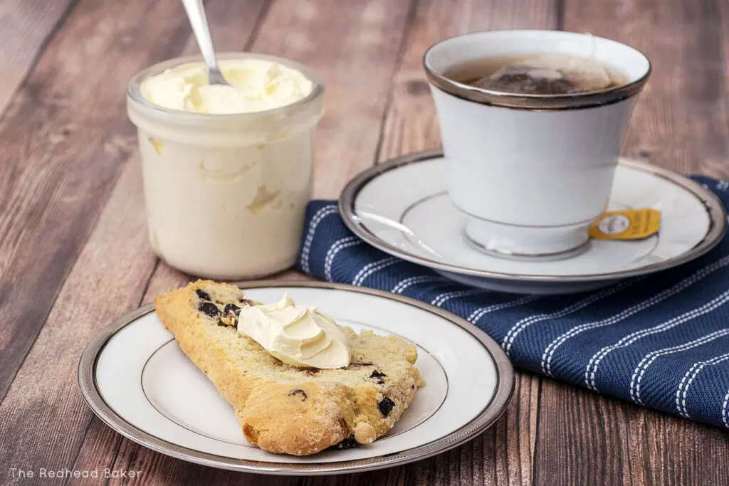 A blueberry scone with a dab of clotted cream in front of a cup of tea and a jar containing more clotted cream