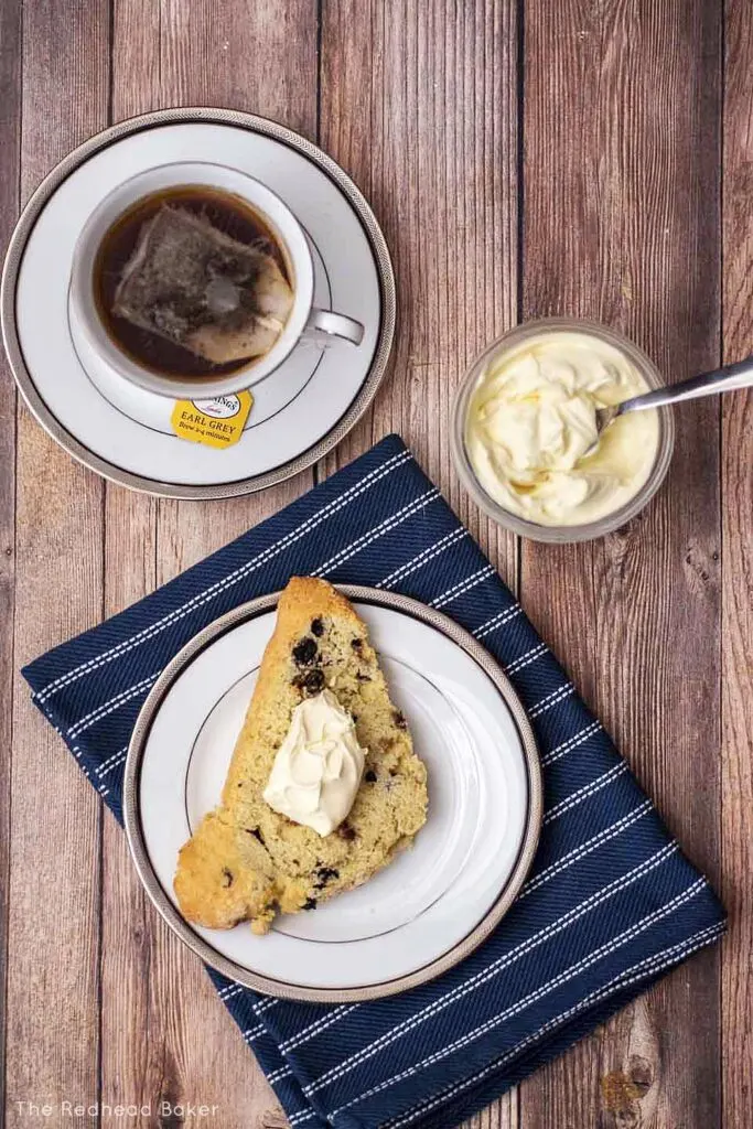 An overhead view of a blueberry scone topped with clotted cream, more cream in a jar and a cup of tea