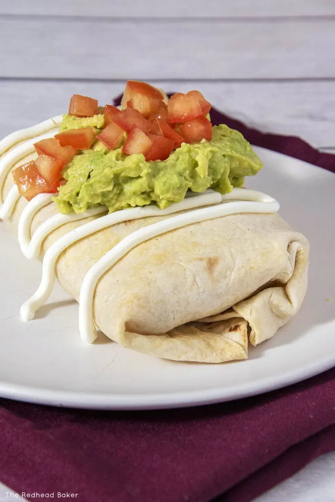 A chicken chimichanga on a white plate topped with sour cream, mashed avocado and diced tomato