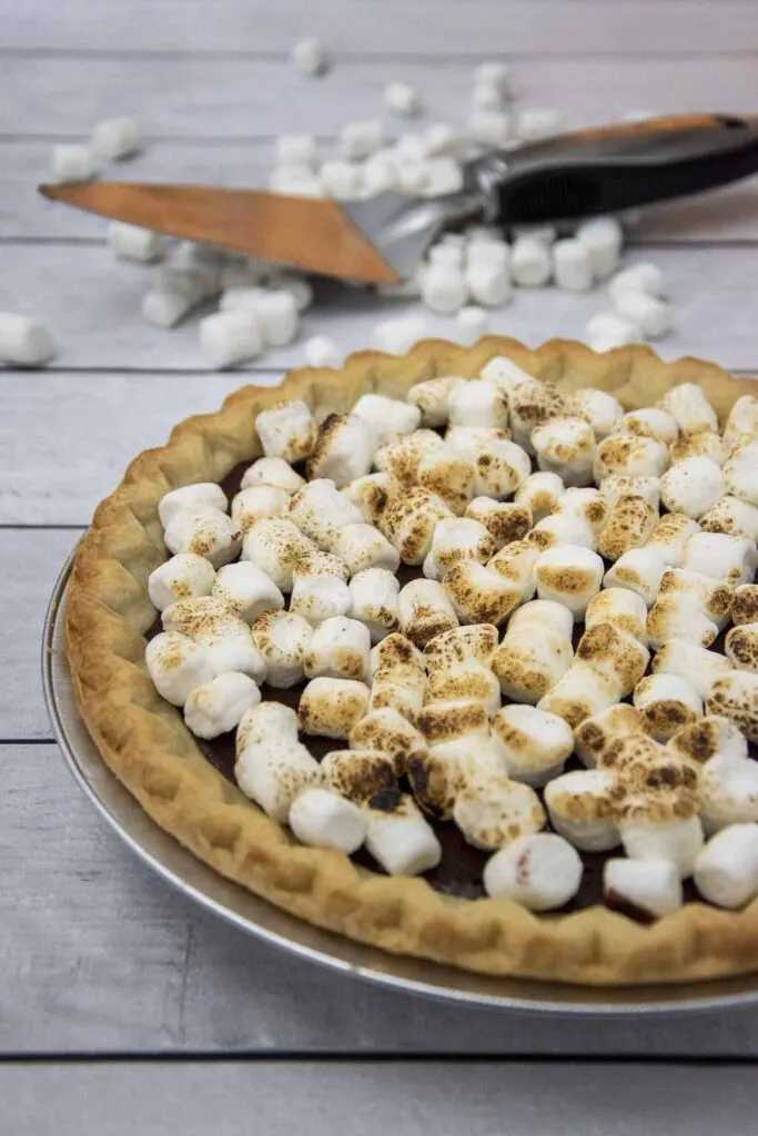 A s'mores pie with toasted marshmallows on top, with mini marshmallows and a pie server in the background