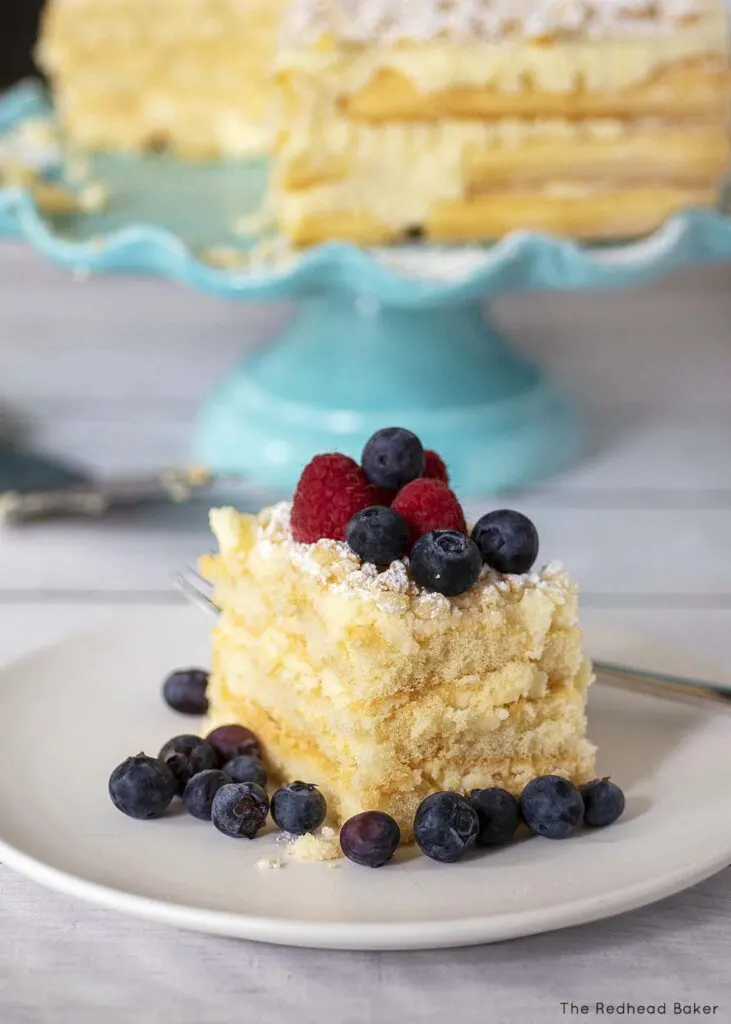 A slice of tiramisu garnished with berries in front of the rest of the tiramisu on a cake stand