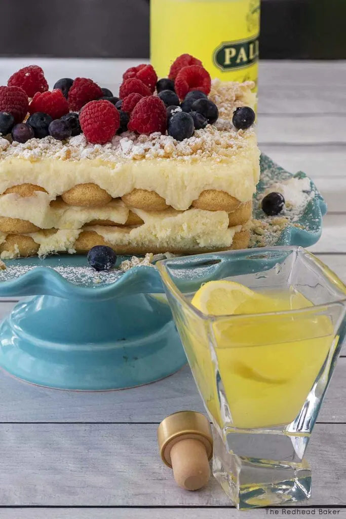 A shot glass of lemoncello in front of a lemon tiramisu on a blue cake stand, int front of a bottle of lemoncello