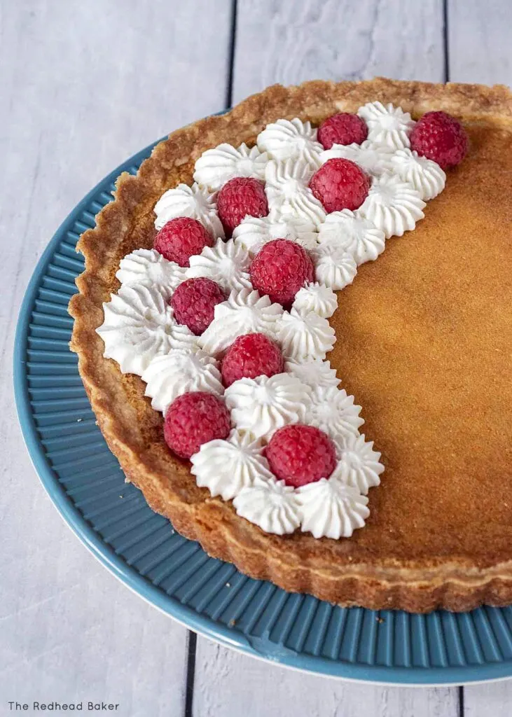 A whole lemon tart garnished with whipped cream and raspberries on a blue plate