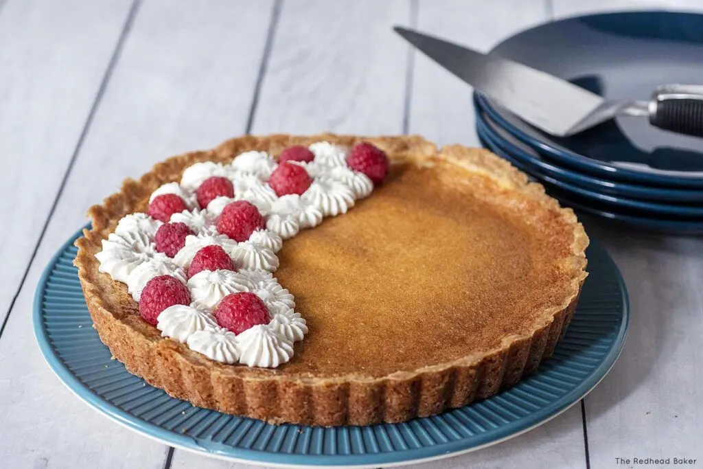 A lemon tart garnished with whipped cream and raspberries on a blue plate in front of a stack of dessert plates and a pie server