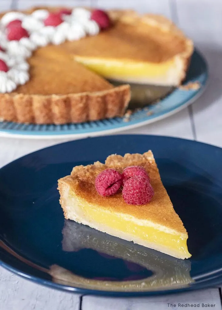 A slice of shortbread lemon tart garnished with raspberries on a blue plate with the rest of the tart in the background