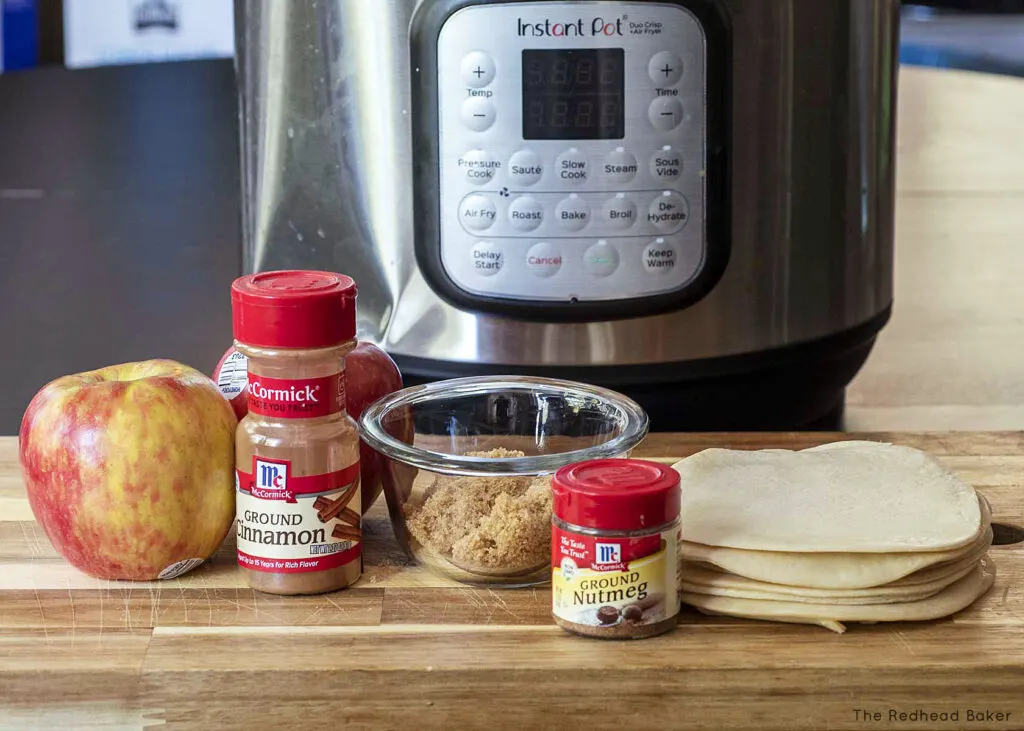 An apple, a bottle of ground cinnamon, a dish of brown sugar, a bottle of ground nutmeg, and a stack of empanada discs on a wooden cutting board in front of an air fryer. 