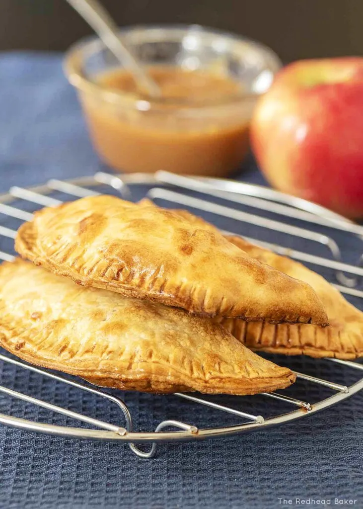 Three air fried apple empanadas on a cooling rack in front of an apple and a dish of caramel sauce.