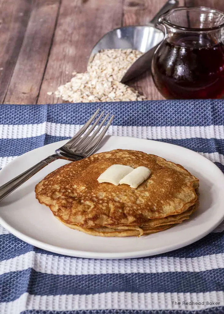 A stack of oat flour pancakes on a white plate, in front of a glass syrup dispenser and a scoop of whole oats