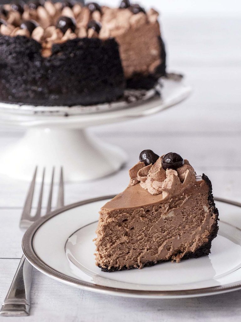 A slice of mocha cheesecake on a white plate in front of the remainder of the cheesecake on a cake stand.