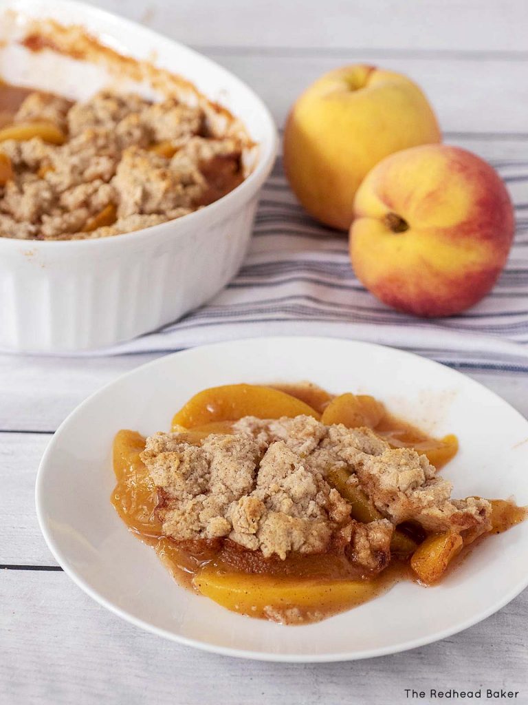 A plate of peach cobbler in front of two fresh peaches and a baking dish