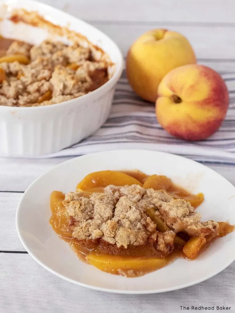 A plate of peach cobbler in front of two fresh peaches and a baking dish