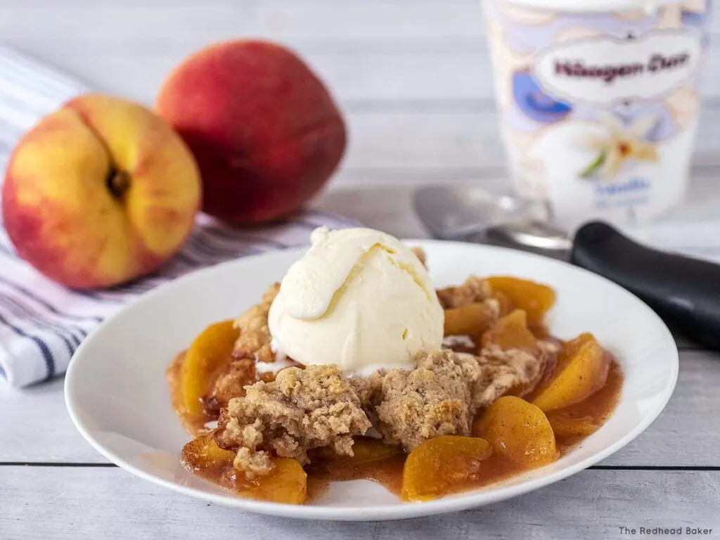 A plate of peach cobbler topped with vanilla ice cream, in front of two peaches and a pint of ice cream.