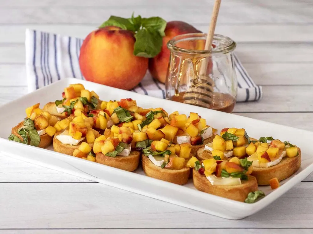 A serving tray of peach bruschetta in front of fresh peaches, basil and a jar of honey.