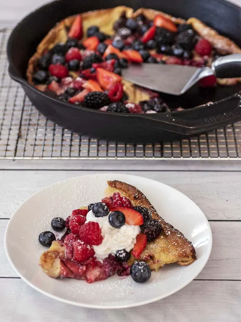 A slice of Dutch baby on a white plate in front of a cast-iron skillet.