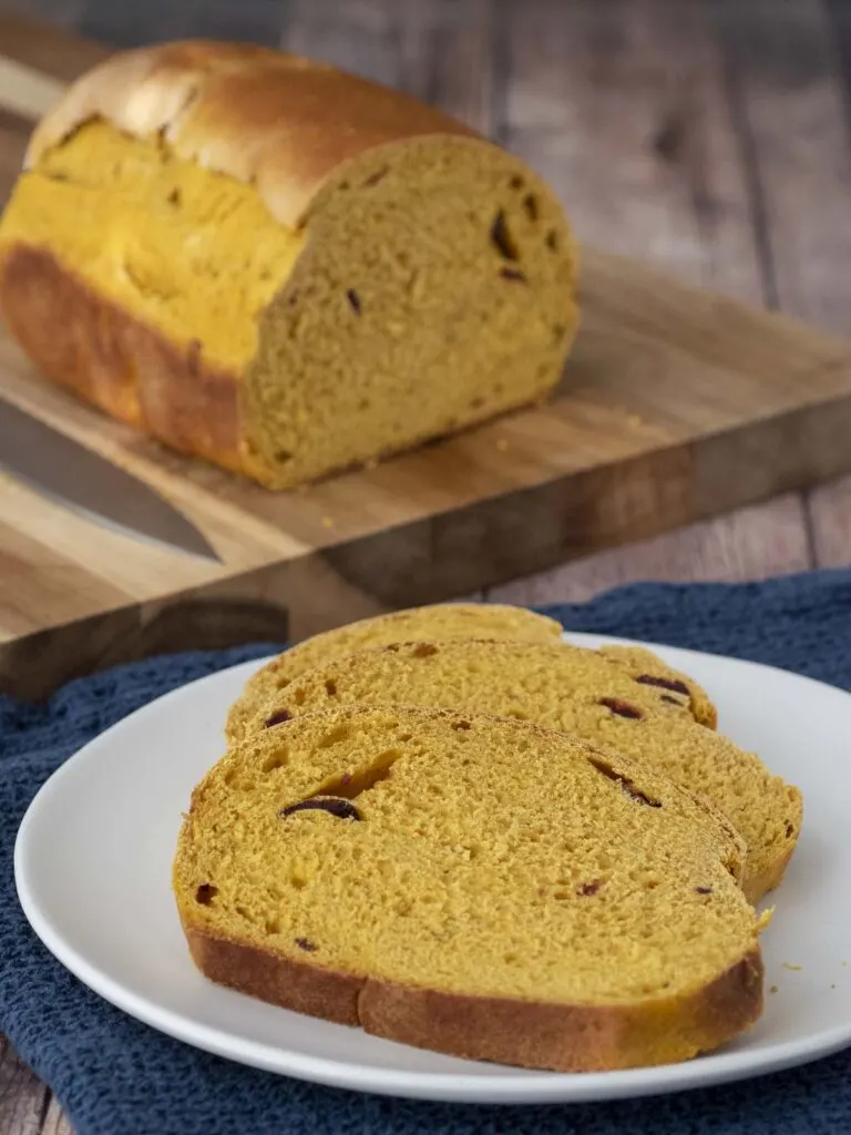 Three slices of pumpkin cranberry yeast bread on a white plate in front of the rest of the loaf.