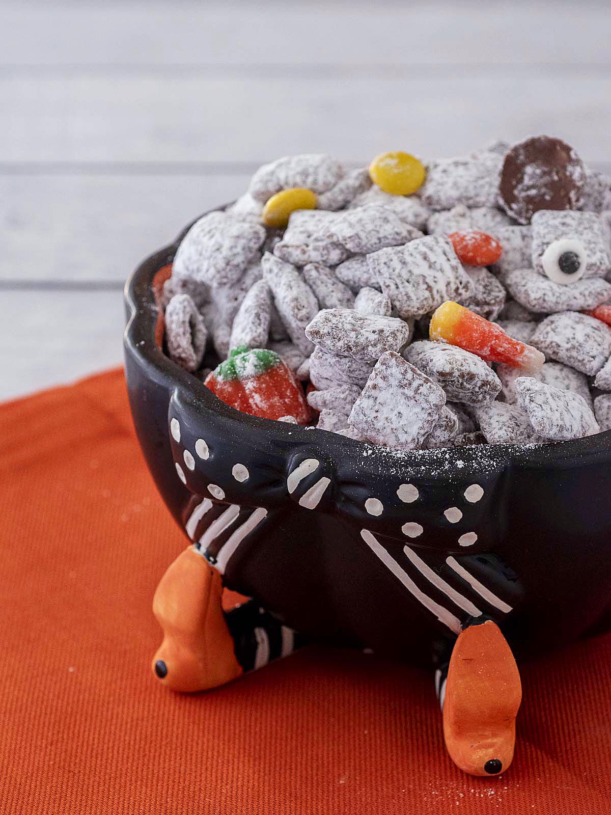 Halloween muddy buddies in a cauldron candy dish on an orange napkin.