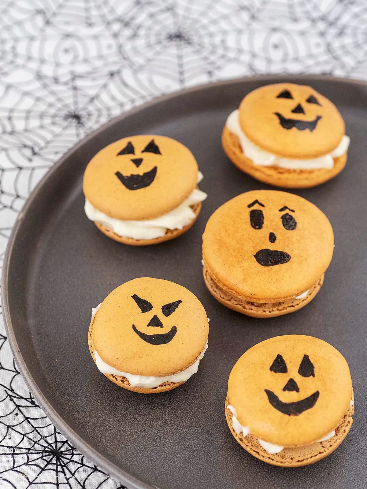 Five jack-o-lantern macarons on a black plate on a spiderweb tablecloth.