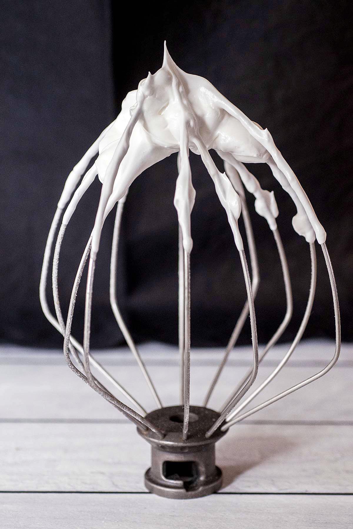 A stand mixer whip attachment with stiff meringue, peaks standing up straight.
