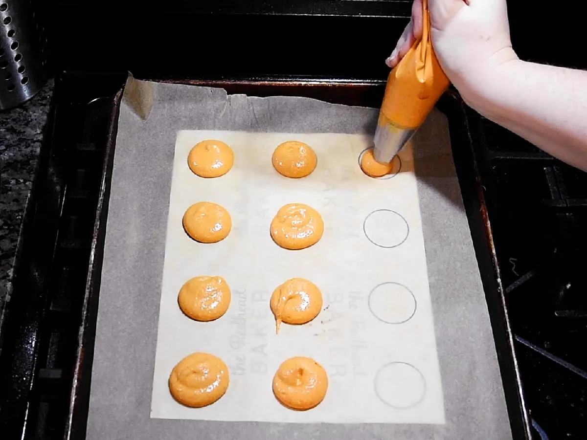 PIping macaron batter onto a parchment-paper-lined cookie sheet with a template underneath.