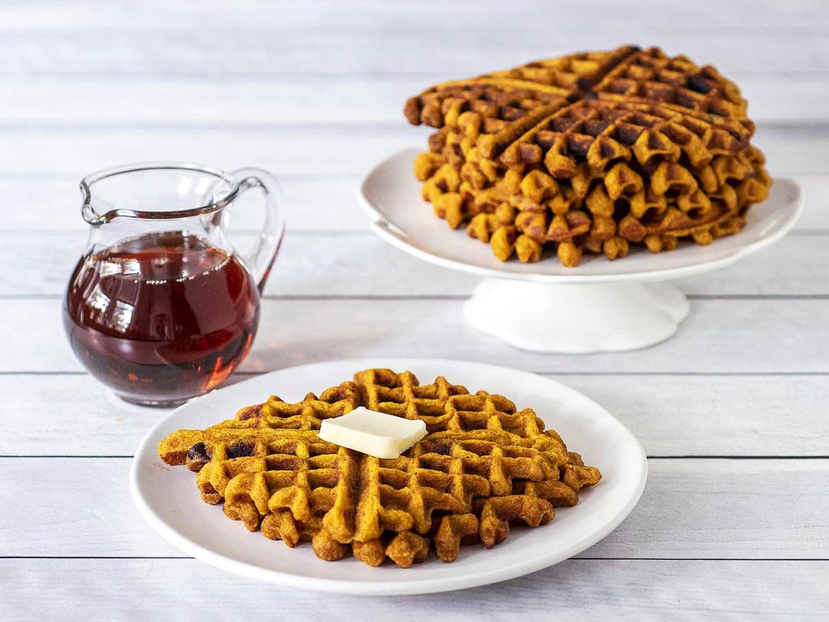 A syrup jar, a white plate with two waffles, and a stack of waffles in the background.