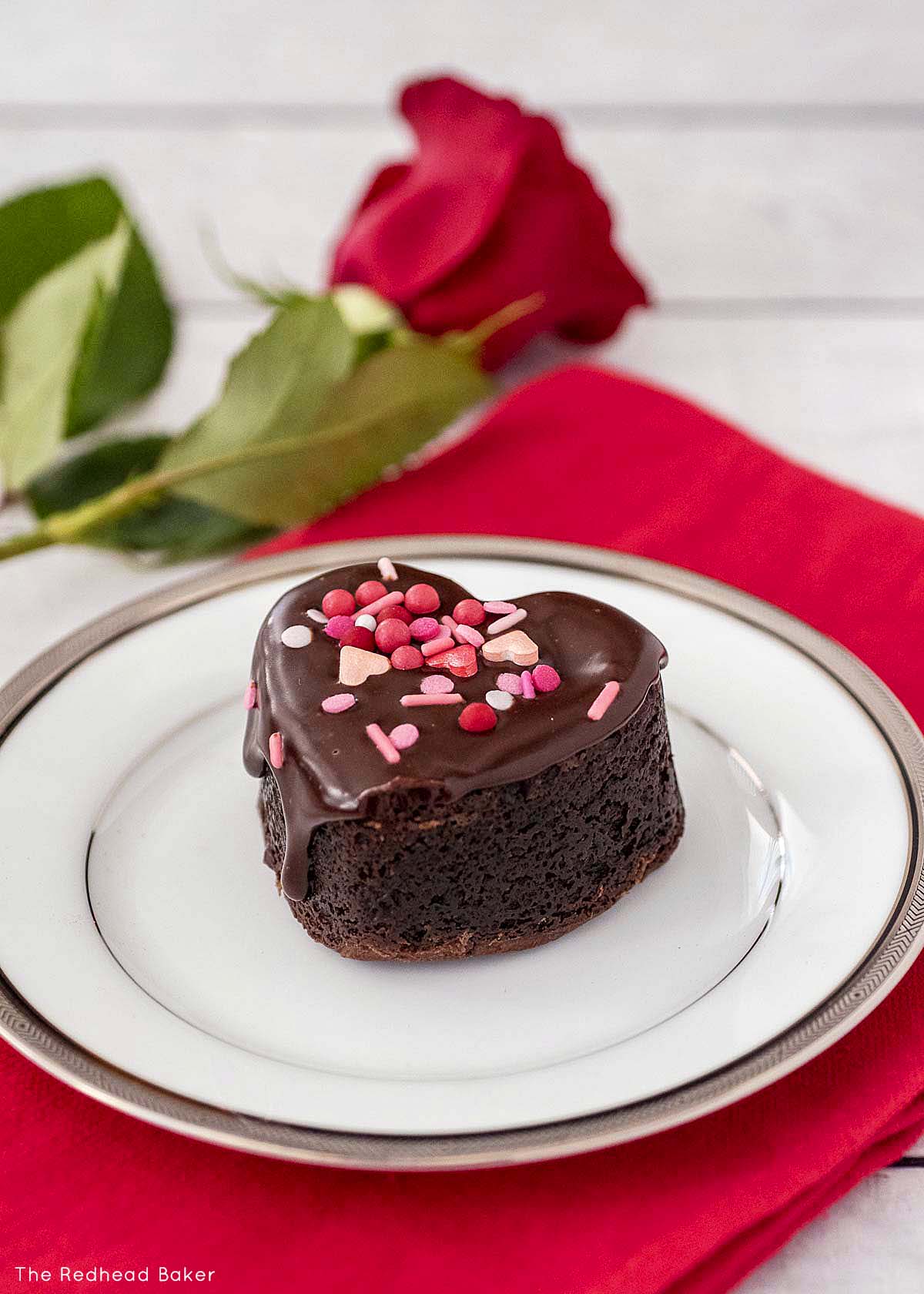 A chocolate-covered brownie heart on a white plate with a red rose.