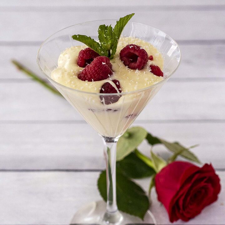 A glass of white chocolate mousse with raspberries in front of a red rose.