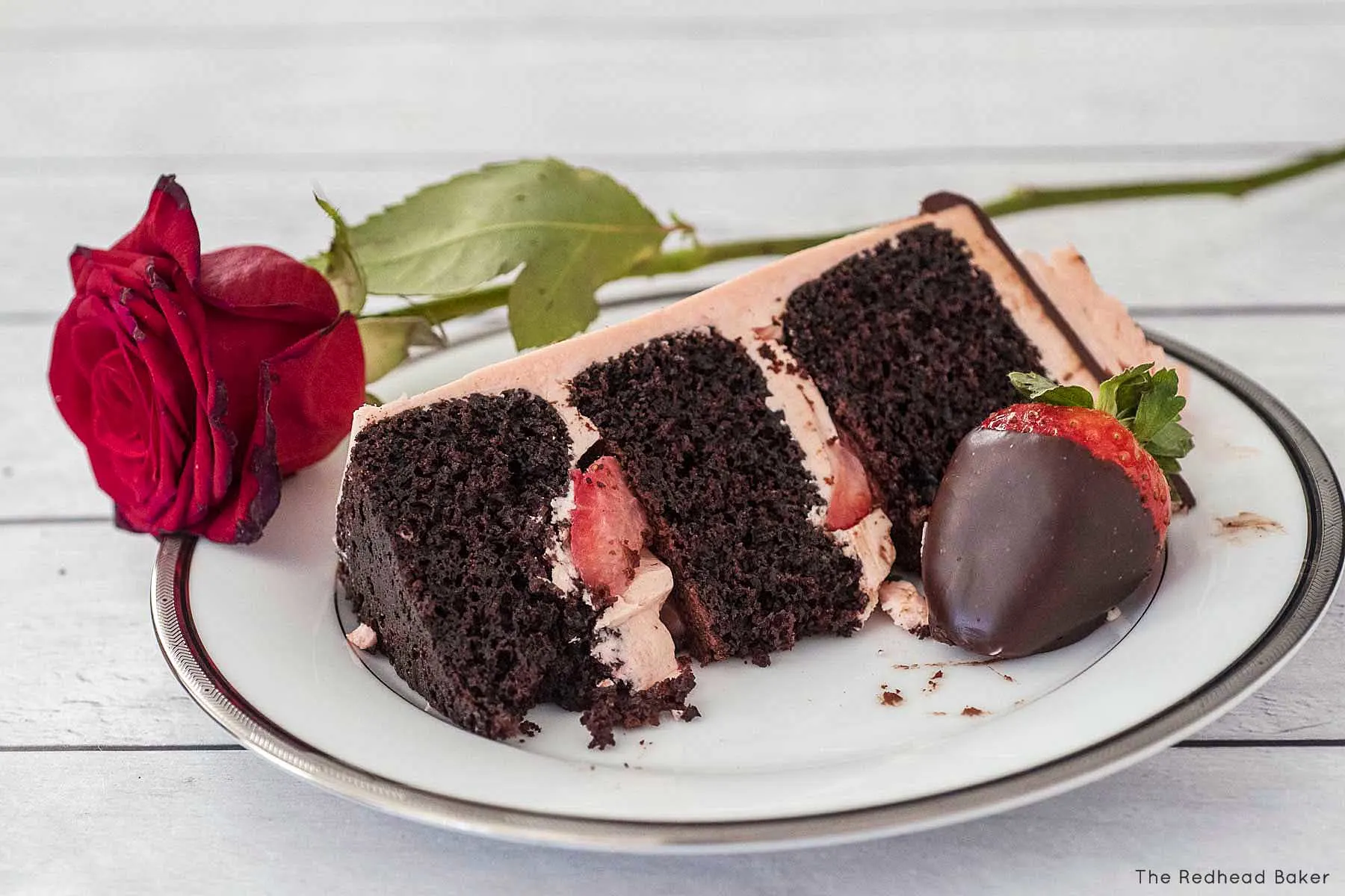 A slice of chocolate layer cake with strawberry frosting and a rose on a white plate.