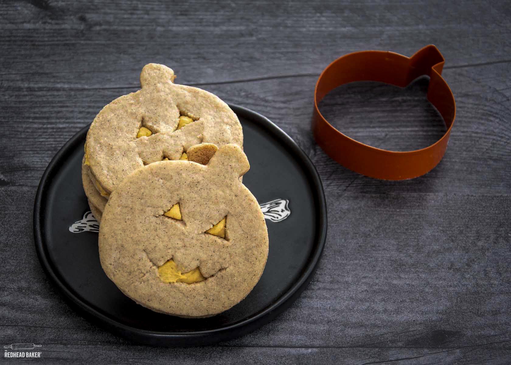 Three jack-o-lantern cookies on a black plate next to a pumpkin-shaped cookie cutter.