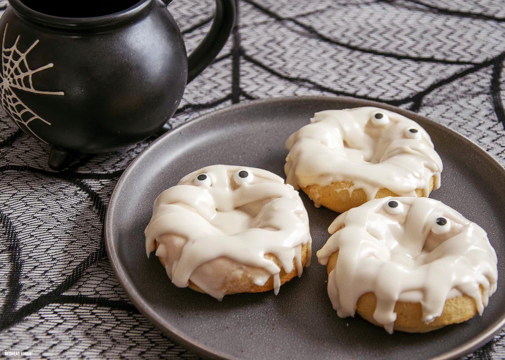 Three baked mummy donuts on a black plate next to a cauldron-shaped coffee mug. 