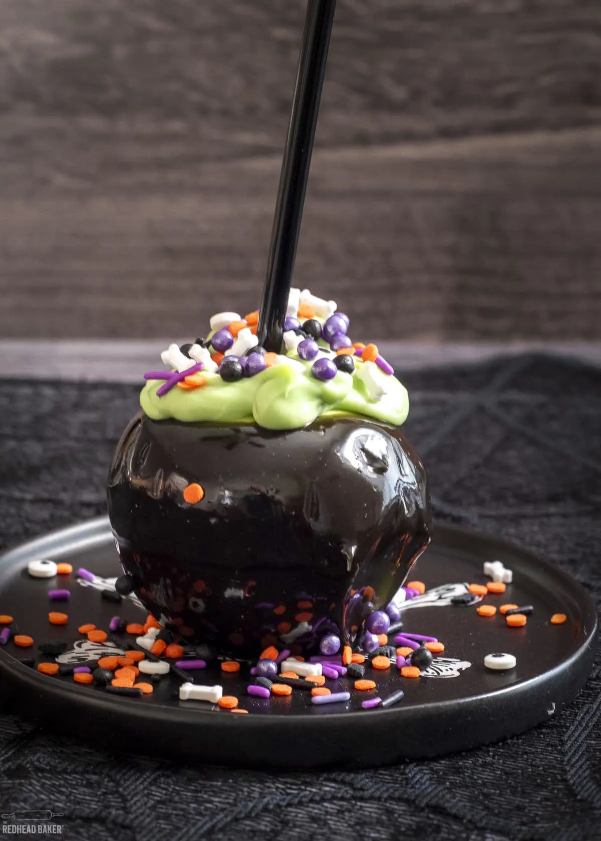A cauldron candy apple on a black plate surrounded by sprinkles.